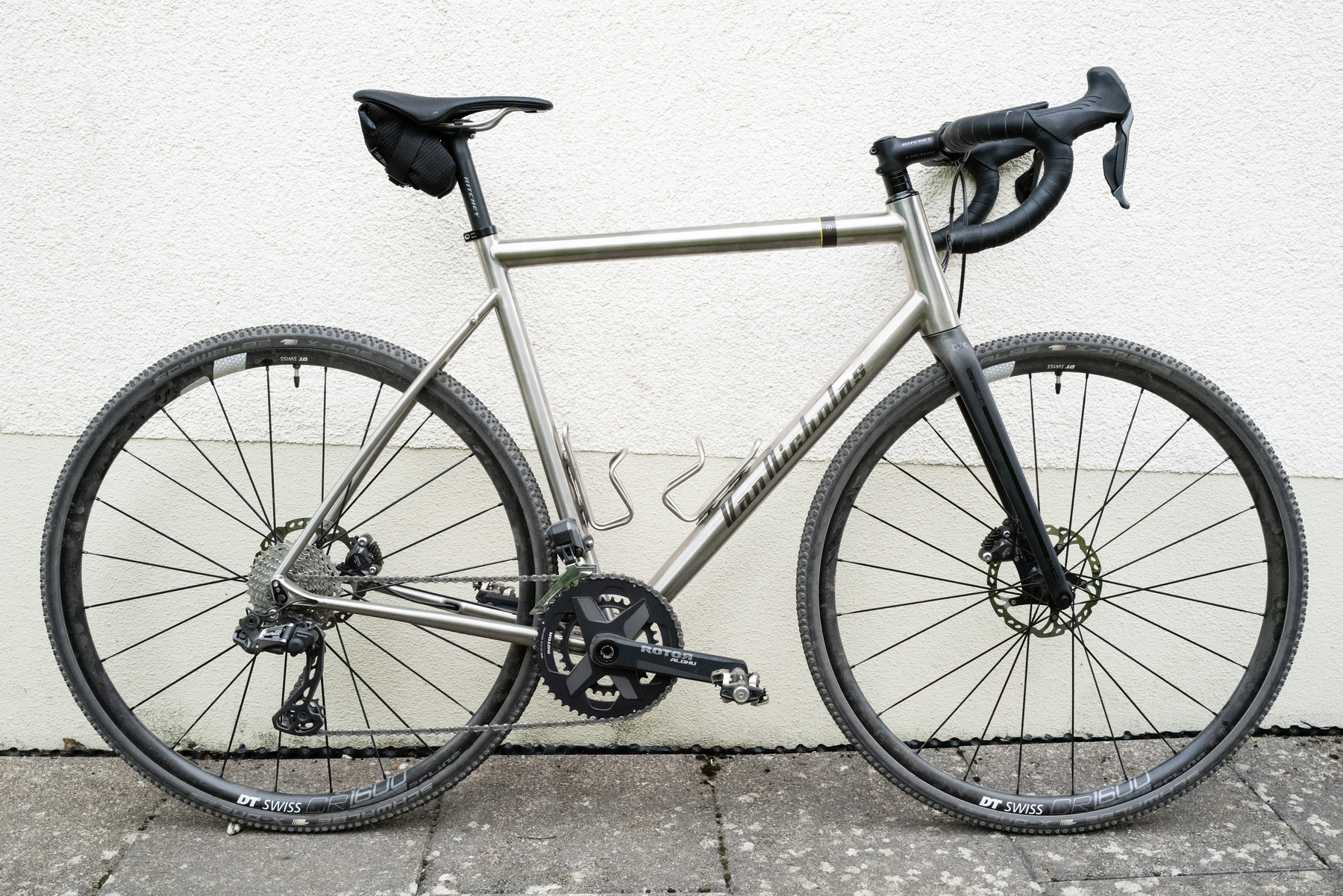 Unpainted titanium-framed bicycle with a black carbon fork, black aluminium rims, knobbly cyclocross tires and a small saddle bag leaning against a house wall. The entire image looks almost monochromatic even though it isn't.